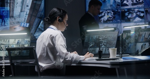 Female security officer works on computer in police monitoring center with CCTV cameras video footage. Male worker controls surveillance cameras with AI face scanning system on big digital screen.