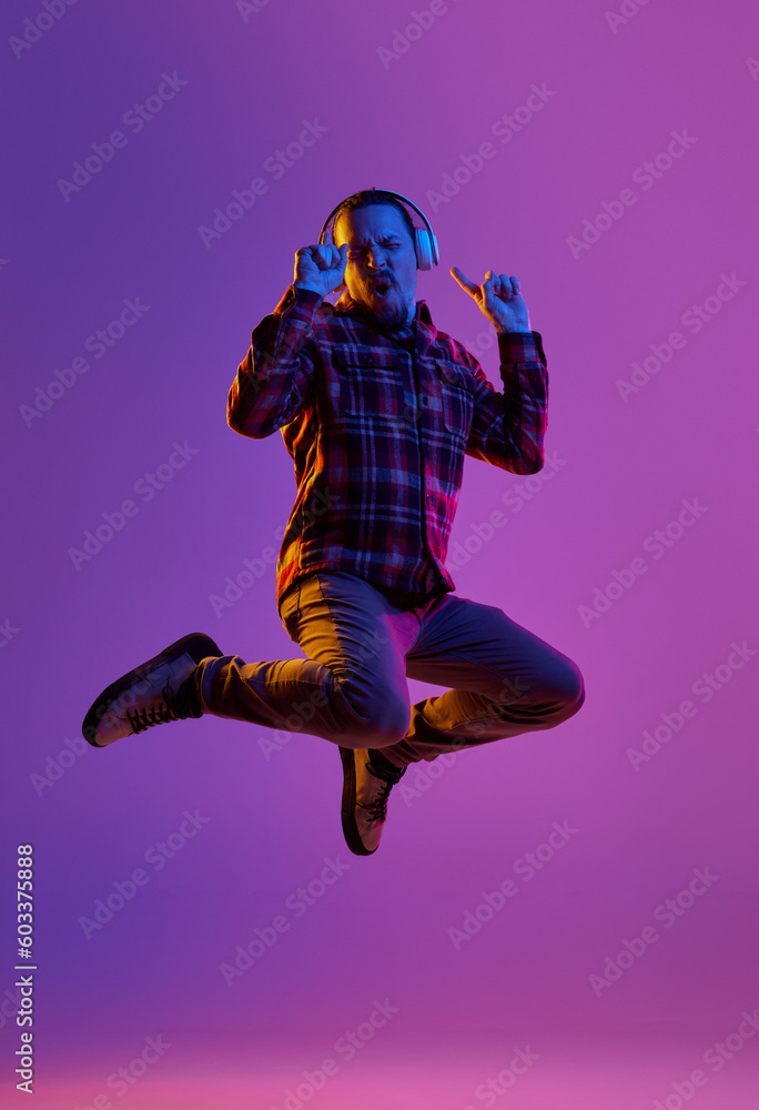 Portrait of emotional man in casual clothes, listening to music in headphones and jumping against purple studio background in neon light. Concept of human emotions, facial expression, lifestyle