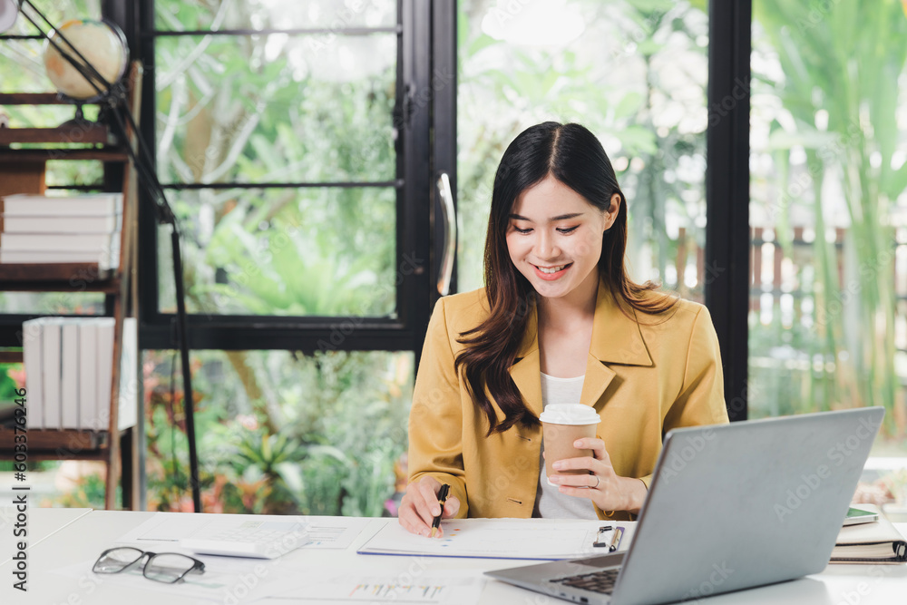 Obraz premium Happy Business woman entrepreneur sit and holding cup of coffee and working with report and laptop at office desk. Portrait of beautiful smiling young businesswoman working at modern work station.
