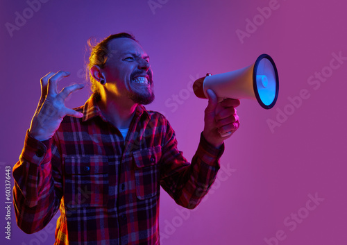 Portrait of angry emotional man shouting in megaphone with annoyance against purple studio background in neon light. News. Propaganda. Concept of human emotions, facial expression, lifestyle