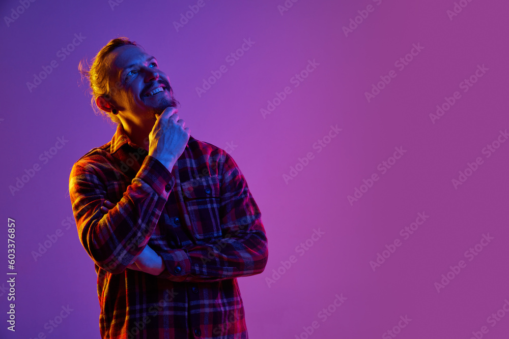 Portrait of happy smiling man with thoughtful face against purple studio background in neon light. Success, ideas. Concept of human emotions, facial expression, lifestyle, business Portrait of happy smiling man with thoughtful face against purple studio background in neon light. Success, ideas. Concept of human emotions, facial expression, lifestyle, business