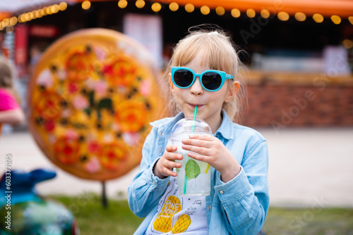 Cute little girl in stylish sunglasses enjoying lemonade drink outdoors.