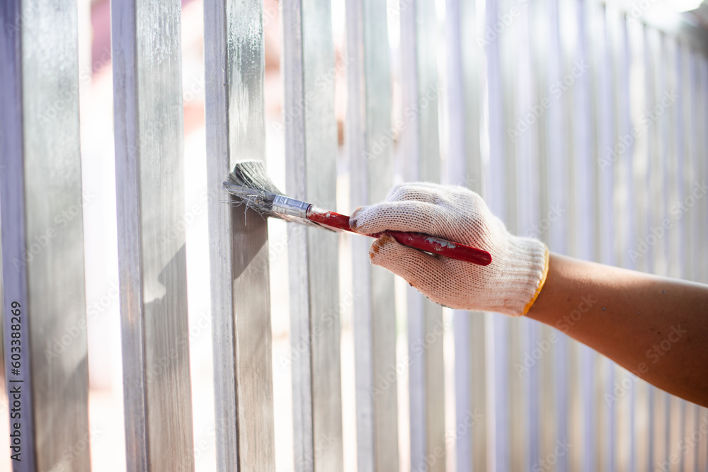Human hand painting a house fence, doing home improvement by refreshing the paint of the steel fence. Close-up  hand wearing glove and holding brush. Fresh wet silver paint.