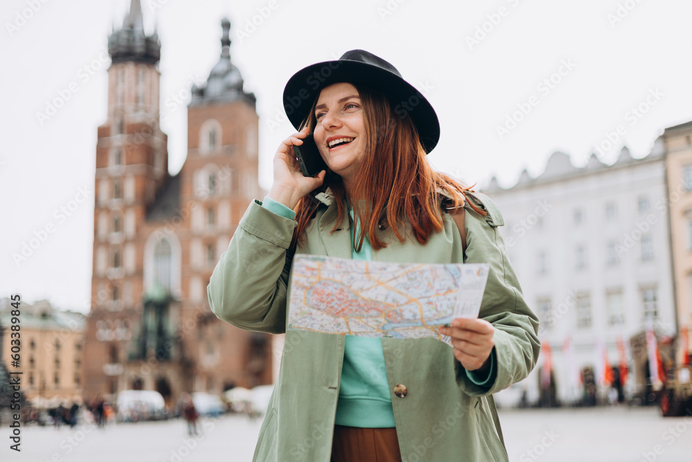 Fototapeta premium Beautiful stylish woman in hat walking on Market Square in Krakow on autumn day and holding mobile phone and map. Phone Communication. Urban lifestyle concept. Check social networks, booking hotel