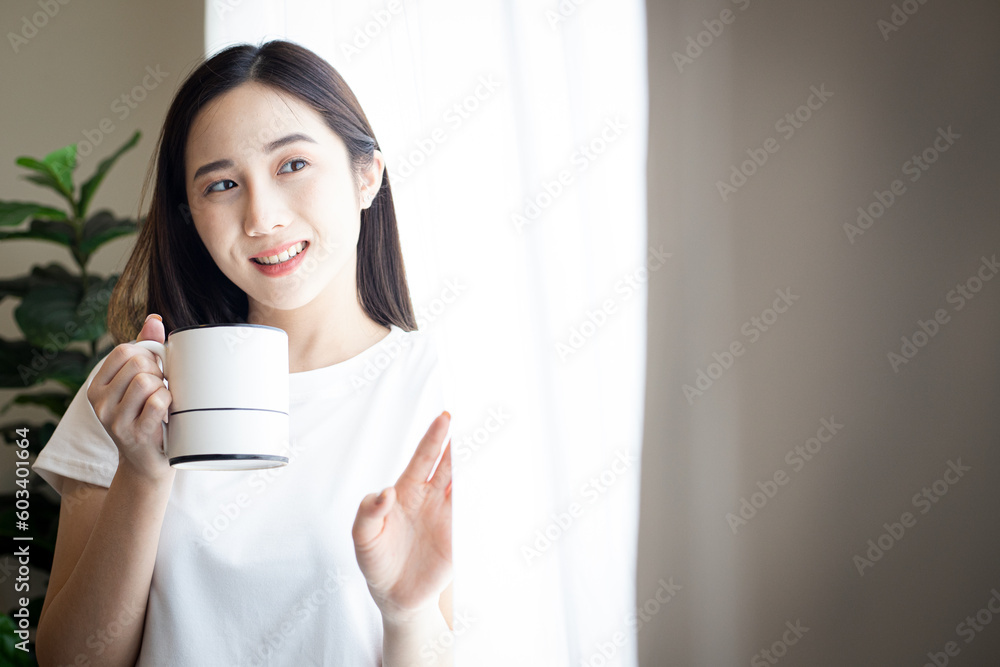 Asian beautiful girl standing near the window with cup of tea or coffee in her hands on the light background in the room Asian beautiful girl standing near the window with cup of tea or coffee in her hands on the light background in the room