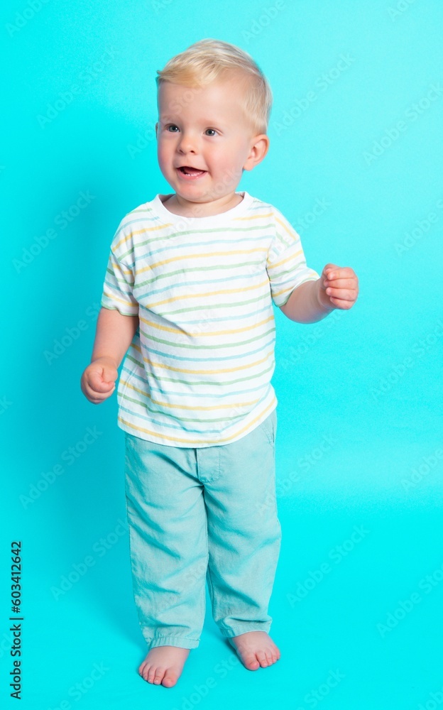 Adorable cute Caucasian blond baby boy striped summer T-shirt trendy clothes set standing barefoot over blue studio background laughing out loud and screaming. Adorable cute Caucasian blond baby boy striped summer T-shirt trendy clothes set standing barefoot over blue studio background laughing out loud and screaming.