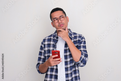Obraz na płótnie Adult Asian man holding mobile phone with suspicious expression