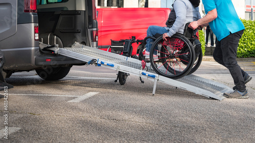 Person on wheelchair with disability using accessible car ramp for transport with help of an assistant driver.