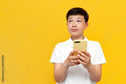pensive little asian boy using smartphone and daydreaming over yellow background, korean child holding mobile