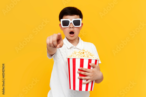 shocked little asian boy with popcorn in 3d glasses pointing forward and wondering on yellow background