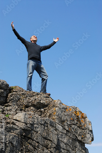 A man standing on a cliff with his arms raised to the blue sky