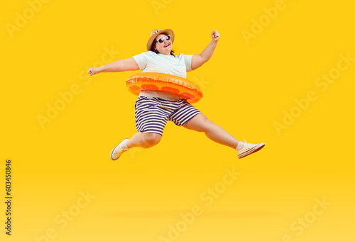 Full body photo of a happy funny fat plus size overweight woman in sunglasses with rubber ring jumping and having fun on studio yellow background. Summer holiday trip and vacation concept. © Studio Romantic