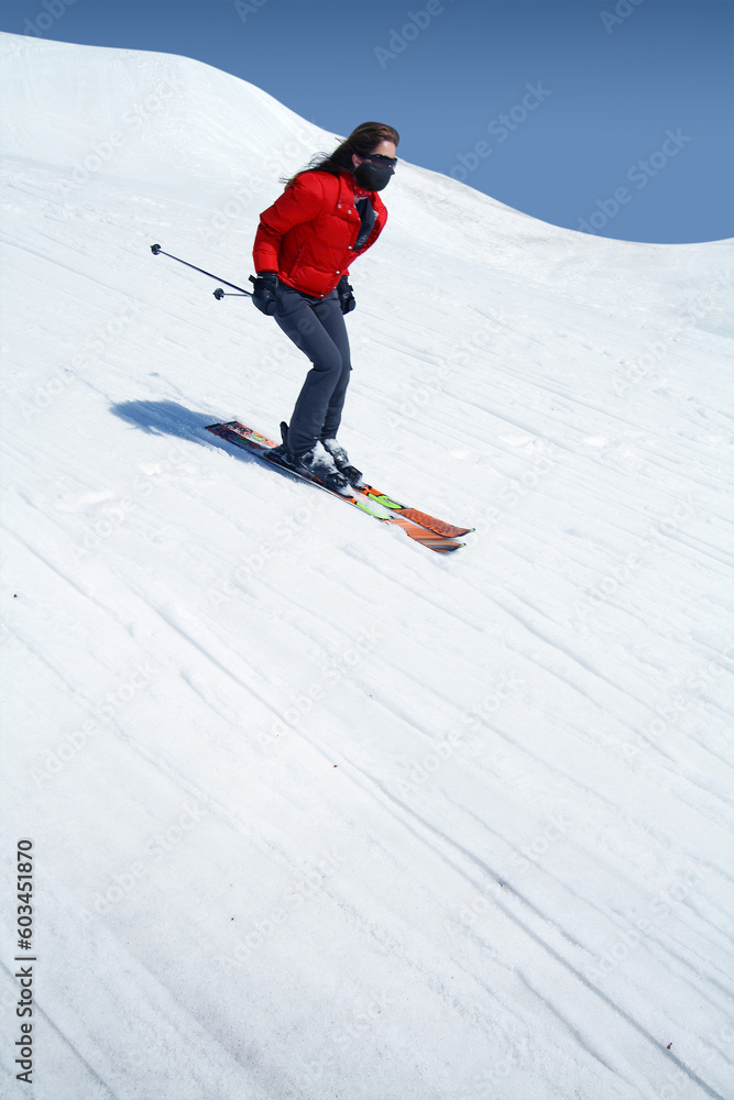 Skier in bright colored outfit skiing down very fast a ski slope in the mountains during winter with blue cloudy skies in the background.