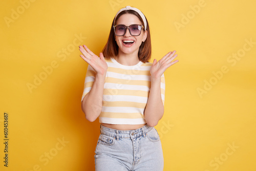 Pleasantly surprised woman wearing striped T-shirt hair band and sunglasses standing isolated over yellow background raised her arms with happy positive expression.