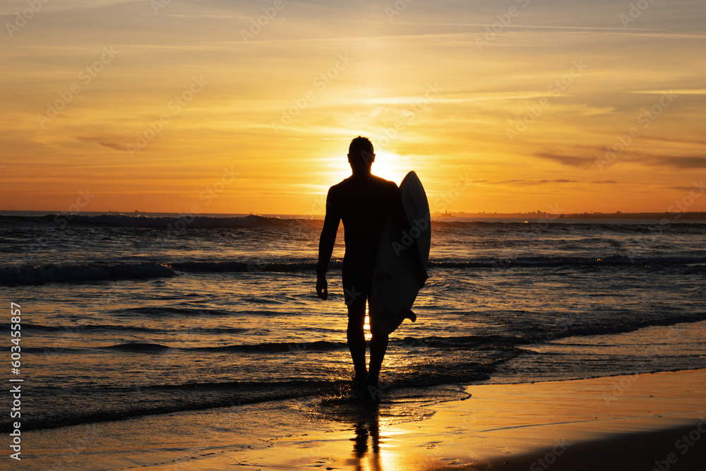 Dark silhouette of a man surfer walking out on the seashore holding a surfboard at sunset Dark silhouette of a man surfer walking out on the seashore holding a surfboard at sunset