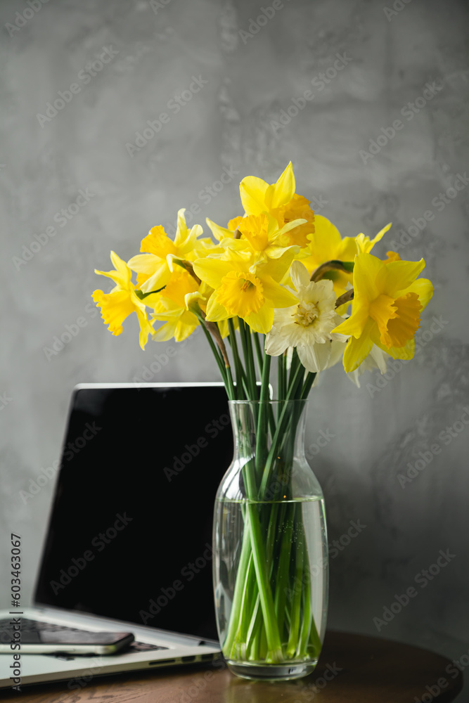 Fototapeta premium Inspired by Spring. Yellow Daffodils flowers in glass vase and Laptop on a table, Grey interior background.