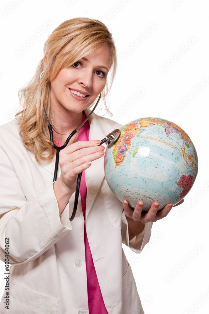 Fototapeta premium Attractive friendly smiling blond lady doctor in white lab coat and pink scrubs holding a stethoscope listening on a globe on white