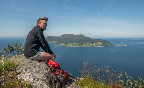 Caucasian tourist with a red backpack sitting on top of Sukkertoppen mountain in Ålesund during summer, enjoying the beautiful view of the surrounding islands and ocean. Norway, day, enjoyment photo