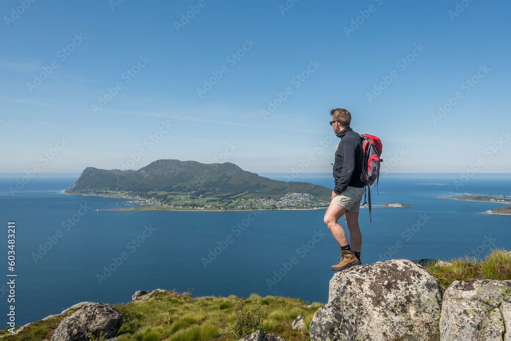 Caucasian tourist with a red backpack standing on top of Sukkertoppen mountain in Ålesund during summer, enjoying the beautiful view of the surrounding islands and ocean. Norway, day, enjoyment