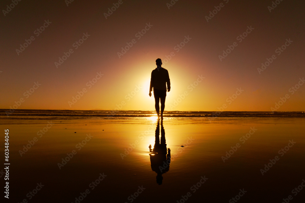 Silhouette of a young man walking on the beach backlit by setting sun and his reflection in the wet sand
