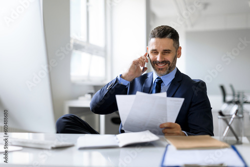 Middle aged businessman talking on cellphone and checking papers, sitting at workplace in office, free space