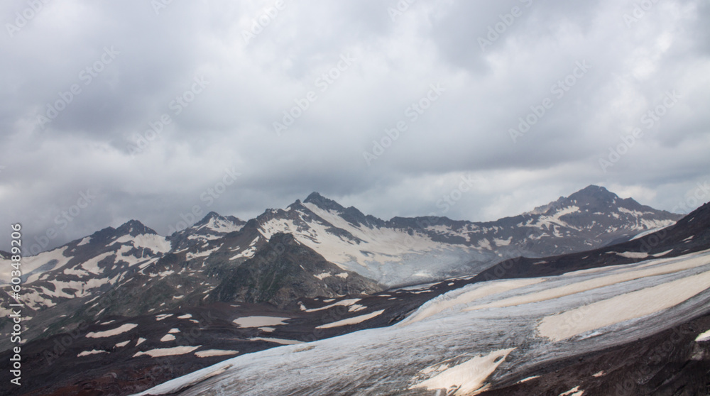 Panoramic landscape - pointed peaks of mountains with white snow and glaciers on the slopes and cloudy sky with dramatic clouds with a blurred horizon in the Elbrus region and copy space