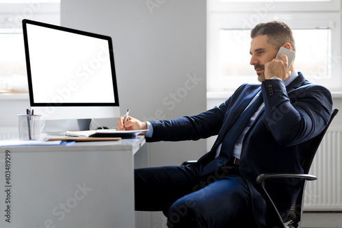Happy man entrepreneur having conversation on cellphone, sitting in front of computer with blank screen, mockup