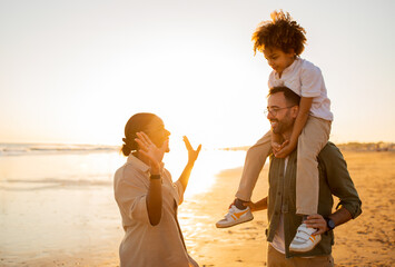 Sand, sea and smiles. Happy young diverse family of three spending free time outdoors, walking and having fun on seaside