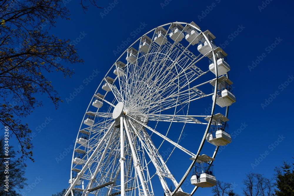 A close up on a white metal ferris wheel located in the middle of a theme park in Poland with numerous compartments for tourists and visitors seen during a sunny summer day in Poland