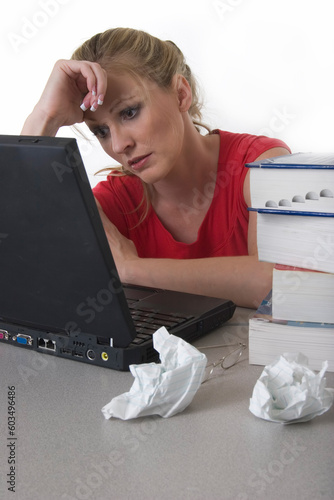 Young woman sitting in front of laptop beside a pile of thick textbooks and crumpled paper with hand on forhead looking stressed photo