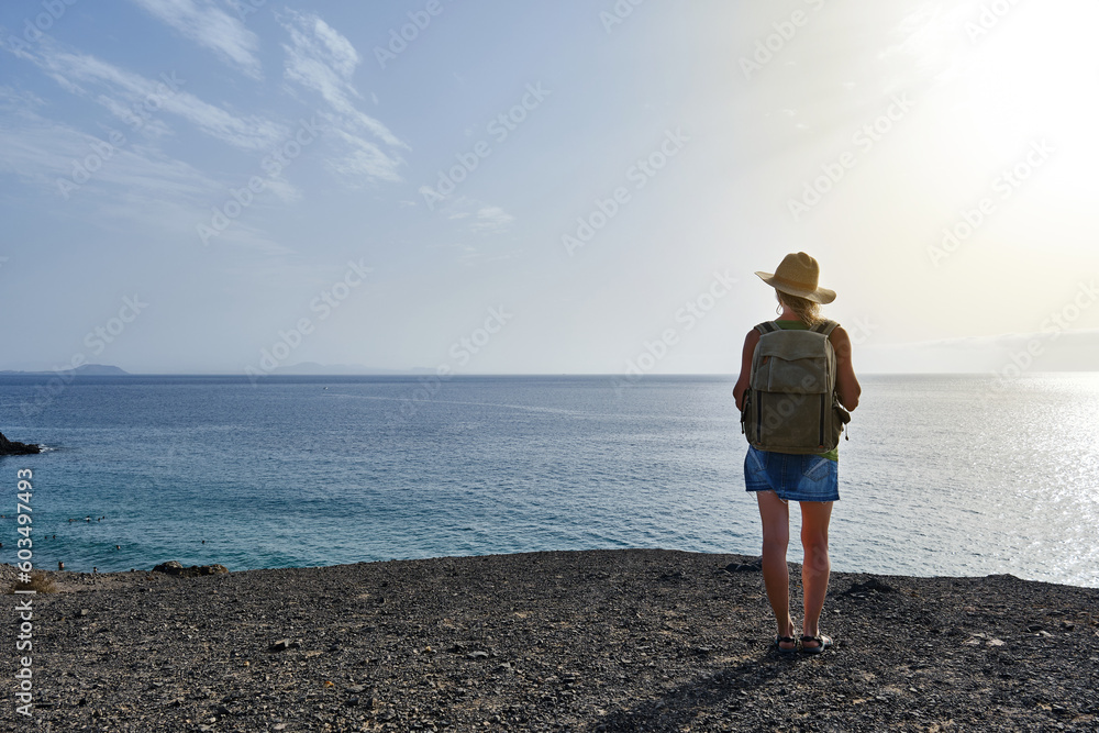 Woman with hat and rucksack in front of a cliff overlooking the sea