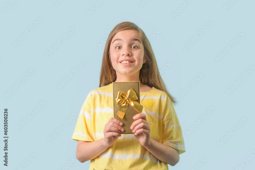 Joyful teenage girl - holds a gift in a gilded package with a bow in her hands. Light blue background Joyful teenage girl - holds a gift in a gilded package with a bow in her hands. Light blue background