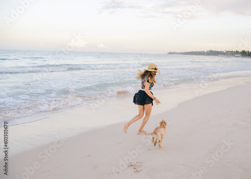 young girl with hat running on the beach with her puppy