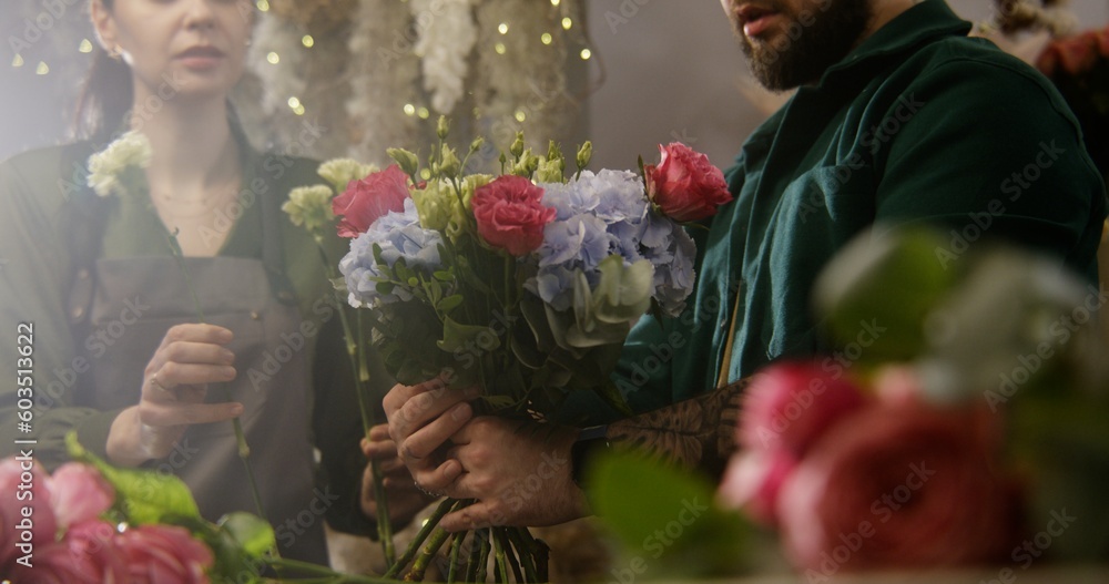 Fototapeta premium Close up shot of professional florist entrepreneur adding fresh flowers to bunch in floral shop. Woman helps colleague to collect beautiful bouquet. Concept of floral business and entrepreneurship.