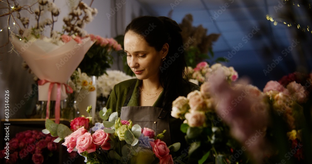 Professional female florist keeps in hands beautiful bouquet, smiles and looks at camera. Vases with plants and fresh flowers stand at background. Retail floral business and entrepreneurship concept.