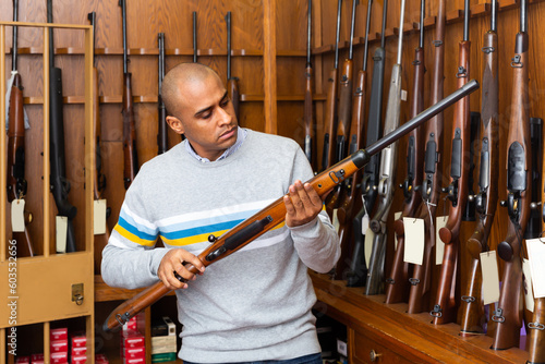 Portrait of focused Latino inspecting sporting shotgun before buying in gunsmith shop