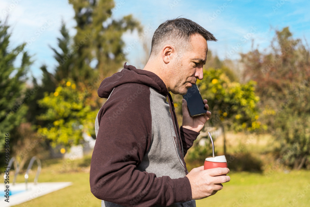 Fototapeta premium Adult man listening to messages on his phone and drinking mate infusion while walking in his garden. Home office concept.