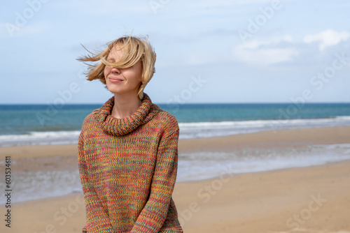 Woman in a sweater on the ocean, sea. Attractive middle-aged woman with windblown hair smiling. Sunny weather, rest, relaxation on vacation. Place for text.