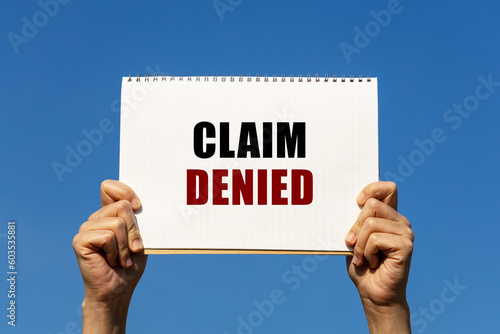 Claim denied text on notebook paper held by 2 hands with isolated blue sky background. This message can be used as business concept about claim denied.