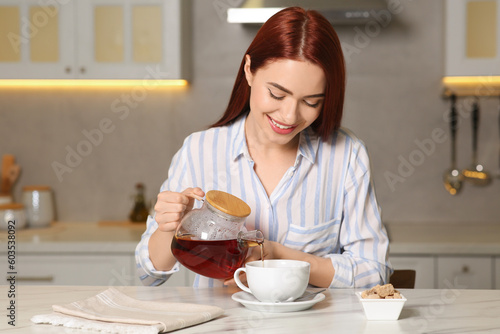 Εκτύπωση καμβά Happy woman with red dyed hair pouring tea from glass teapot into cup at white t