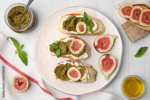Tasty bruschettas with cream cheese, pesto sauce, figs and fresh basil on white tiled table, flat lay