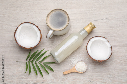 Bottle of delicious syrup, halves of coconut, flakes, cup of coffee and green leaves on white wooden table, flat lay