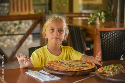 An american curly blonde boy eating a pizza in italian restaurant. Portrait of child with long blonde hair and a yellow T-shirt eats junk food in a cafe. Family holiday in Italia. looks at the camera