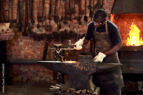 Fototapeta Naklejka Na Ścianę i Meble -  Blacksmith, worker and metal forge in workshop and manual industry working on hot steel with hammer, sparks or fire. Black man, welding or iron tools manufacturing or expert, trade and dark workspace