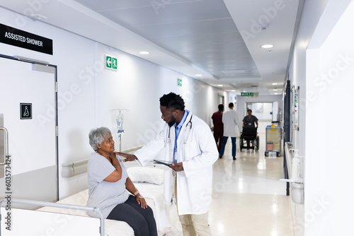 African american male doctor with tablet talking to diverse senior female patient in busy corridor