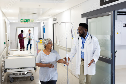 African american male doctor talking to diverse senior female patient with drip walking in corridor