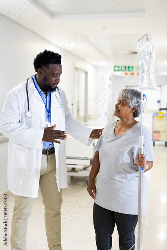 African american male doctor talking to diverse senior female patient with drip standing in corridor