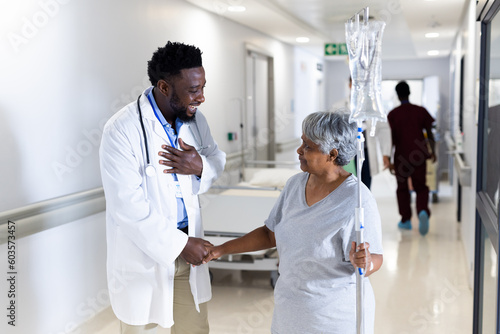 Biracial senior female patient with drip shaking hand of happy diverse male doctor in corridor