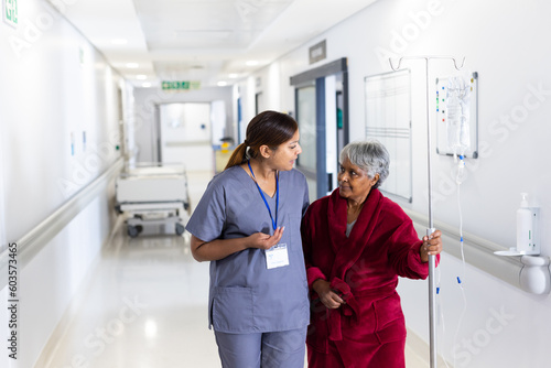 Asian female doctor talking to diverse senior female patient with drip walking in corridor
