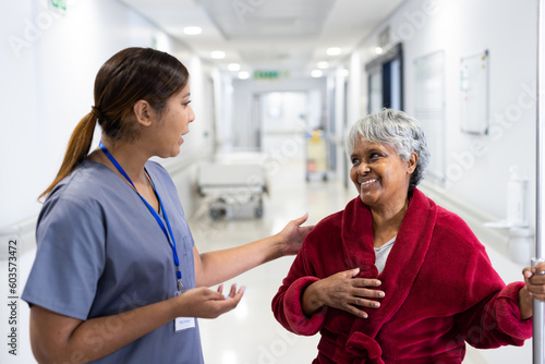 Asian female doctor talking to smiling diverse senior female patient in hospital corridor